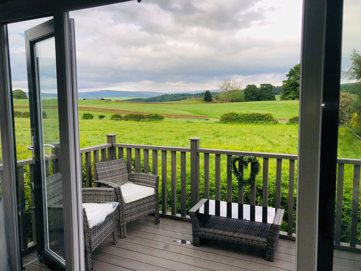 A spacious outdoor veranda features two wicker chairs with white cushions and a glass-top table. The area overlooks expansive green fields and rolling hills, creating a serene countryside view framed by large glass doors.