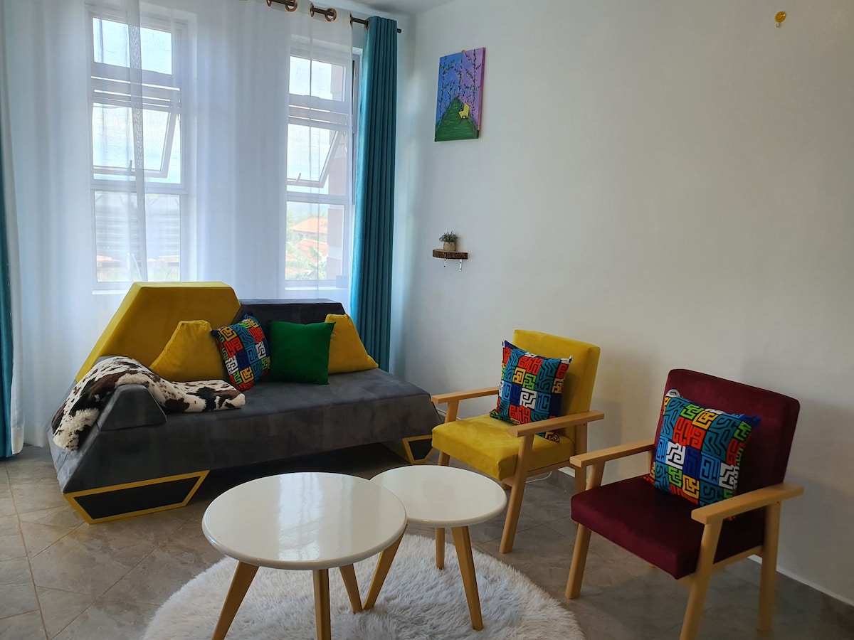 A cozy living area features a modern gray sofa with colorful cushions, complemented by two armchairs in yellow and burgundy. A round coffee table sits on a soft rug, while natural light streams through sheer curtains covering the windows.