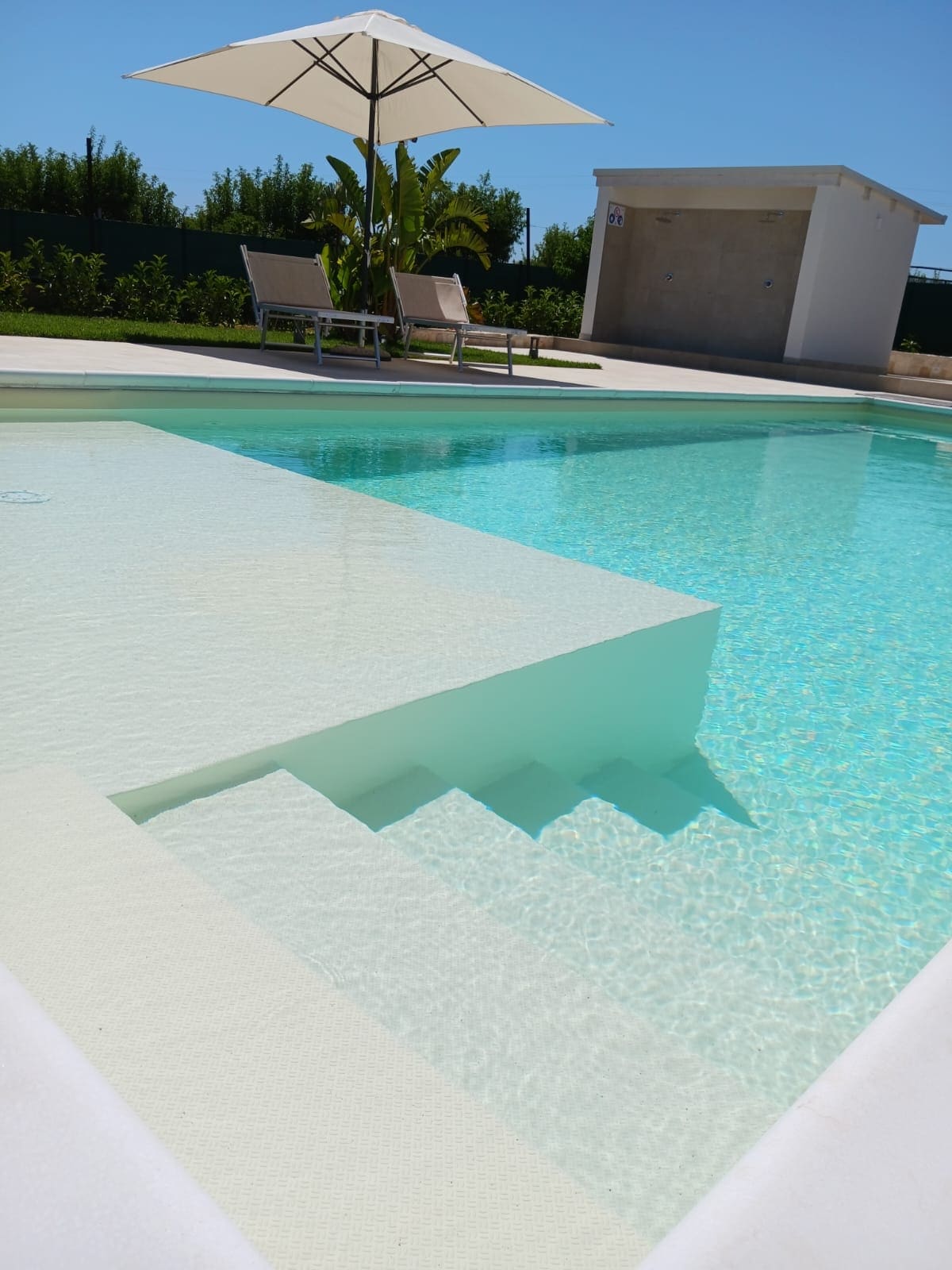 A private swimming pool is featured, with steps leading into the clear water. A sun umbrella provides shade for a pair of lounge chairs positioned nearby. Lush greenery surrounds the pool area, complemented by a well-maintained exterior space.