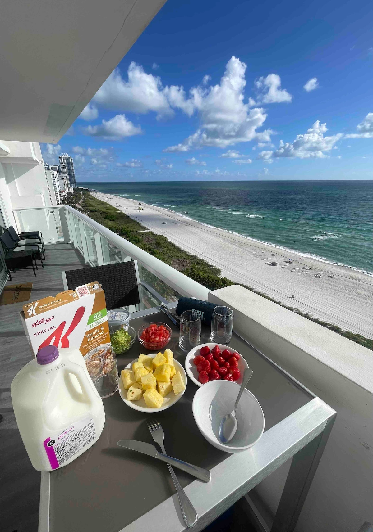 A balcony table is set with a selection of breakfast items, including fresh fruits like pineapple, strawberries, and tomatoes, accompanied by a box of cereal and milk. The view features a sandy beach and the ocean under a bright sky with scattered clouds.