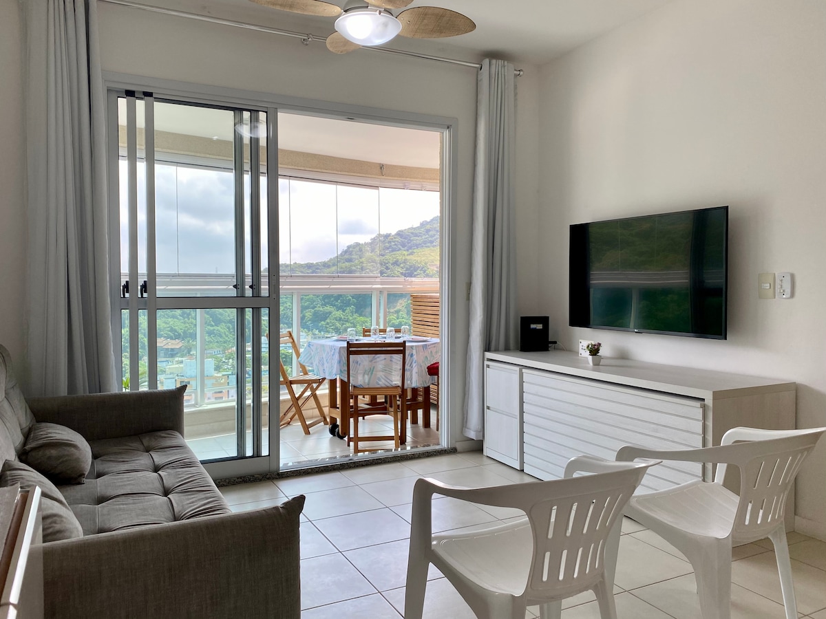 A bright living area features a comfortable sofa and two white chairs facing a TV mounted on a white media console. Large glass doors open to a balcony with a dining table, showcasing views of surrounding hills and greenery.