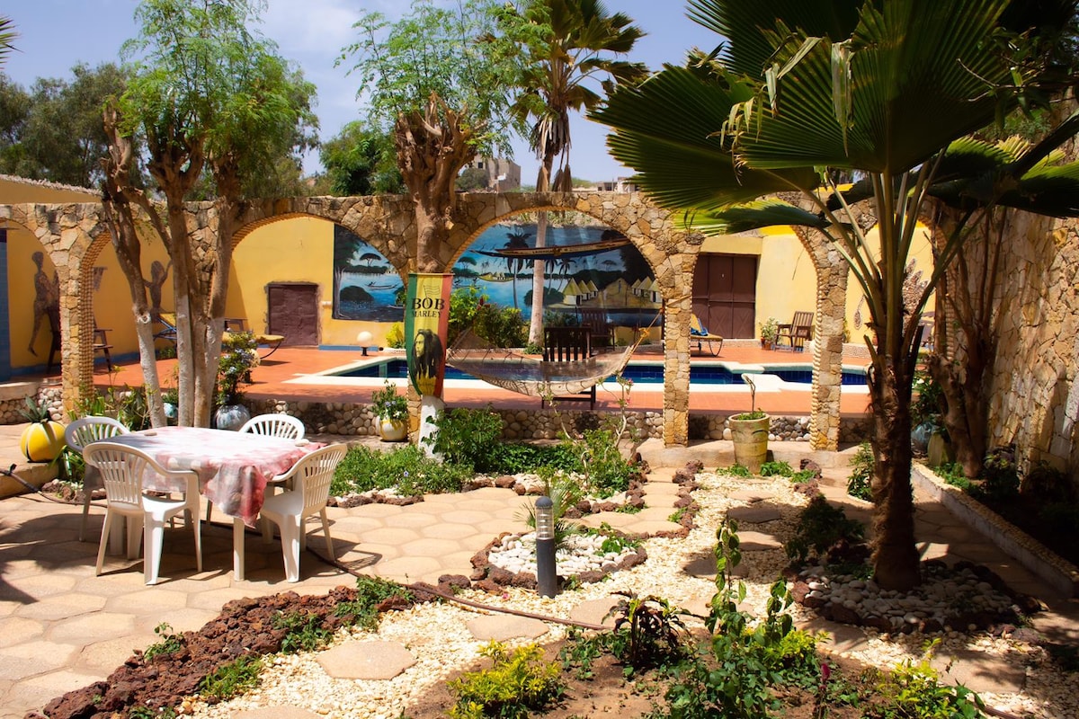 A garden area features a circular stone seating arrangement surrounded by greenery. A swimming pool is visible in the background, with vibrant murals adorning the walls. Tall palm trees provide shade, and decorative plants enhance the serene environment.