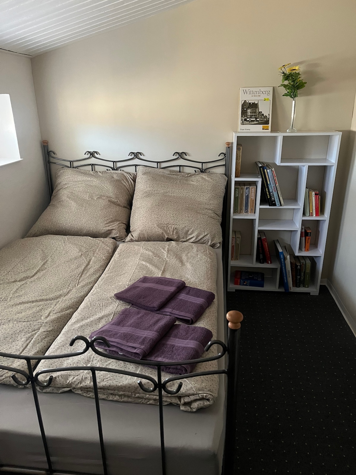 A cozy sleeping area features a double bed with soft bed linens and two purple towels neatly arranged on top. A small bookshelf holds various books and a vase with a flower. Natural light enters through the window beside the bed.
