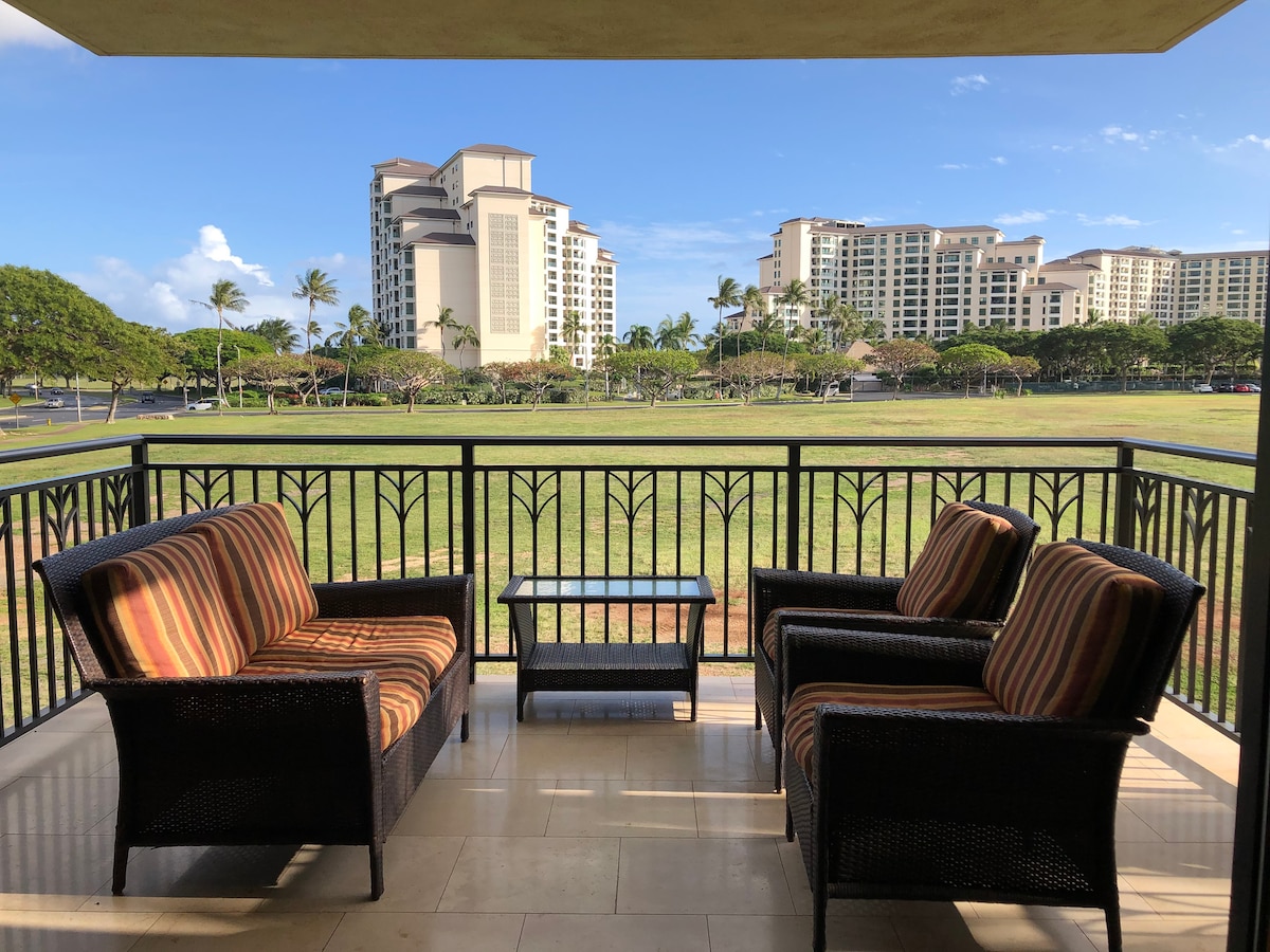 A private balcony is furnished with two spacious wicker chairs featuring striped cushions and a small glass table. The view extends over a grassy open field, framed by palm trees and a backdrop of resort-style buildings under a clear blue sky.