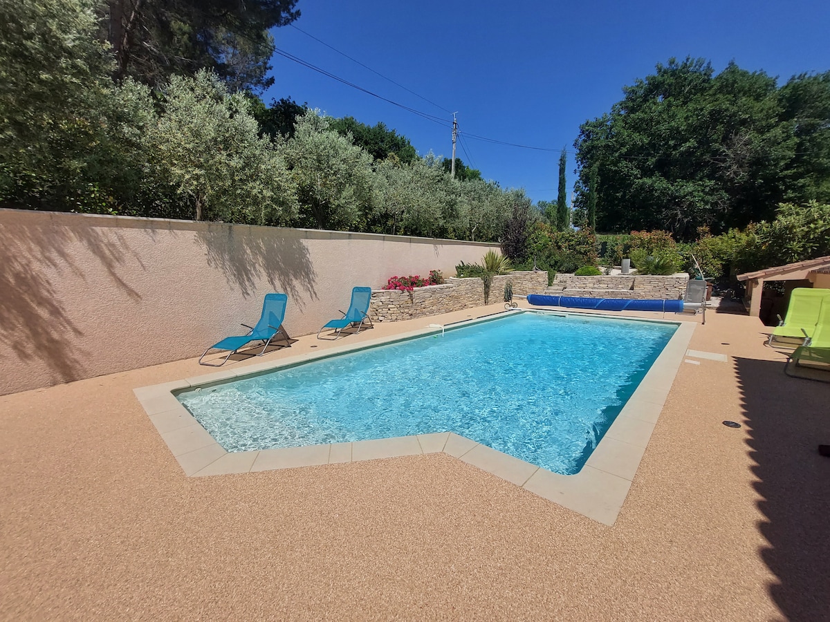 A clear swimming pool is framed by a light-tiled deck, featuring two blue lounge chairs positioned nearby. The surrounding area is decorated with lush greenery and colorful flowers, creating a serene outdoor space. The sky is bright and blue, enhancing the inviting atmosphere.