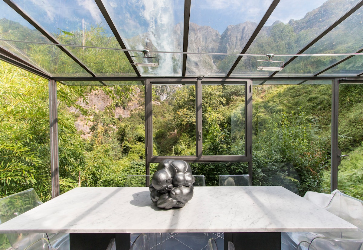 A glass-enclosed dining area features a marble table at its center, adorned with a sleek black sculpture. Verdant foliage and the mountainous landscape are visible through the glass walls, creating a serene connection to the outdoors.