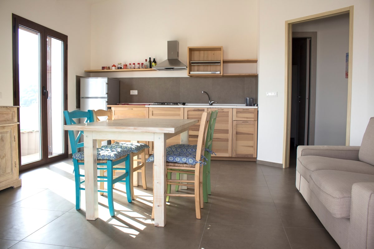 A modern kitchen and dining area is presented with light wood furniture. A table with blue and green chairs is centrally located, while a functional kitchen with a stove and refrigerator is visible in the background. Soft natural light brightens the spacious layout.