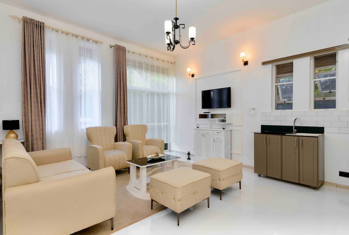 A contemporary living space is showcased, featuring two armchairs and a sofa in soft beige tones. A sleek coffee table sits in the center, complemented by two matching ottomans. Natural light floods in through large windows, while a wall-mounted television and a small kitchenette add functionality.
