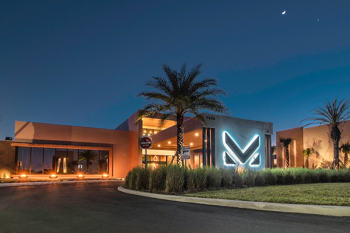 The modern exterior of the townhouse features a sleek facade illuminated by soft lighting. Palm trees frame the entrance, highlighting the welcoming atmosphere of the community. A spacious driveway leads to the main entrance, which is accentuated by the contemporary illuminated logo.