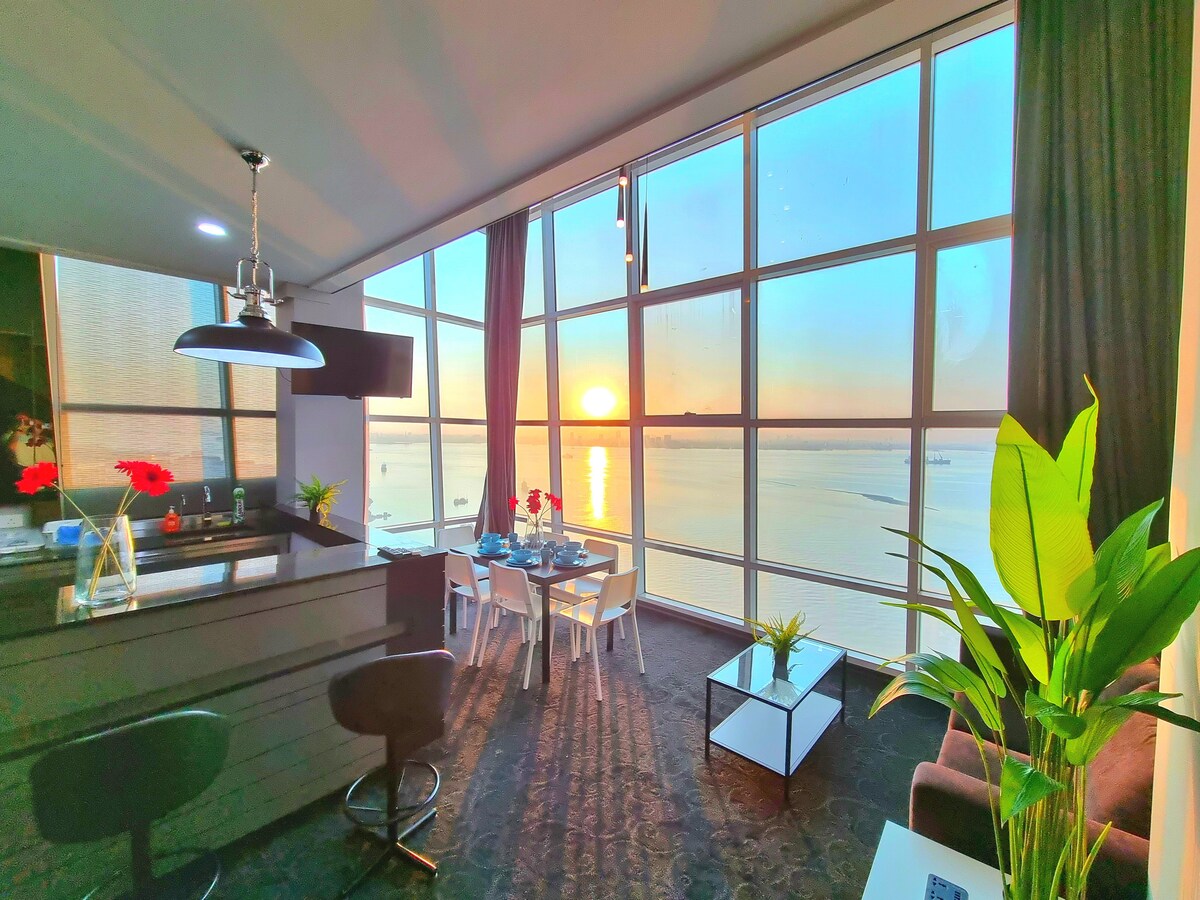 A spacious living area is illuminated by the warm glow of a sunrise, with large windows showcasing a panoramic seaview. A dining table set for four is positioned near the glass, alongside a minimalist coffee table and indoor plants, creating a serene environment.