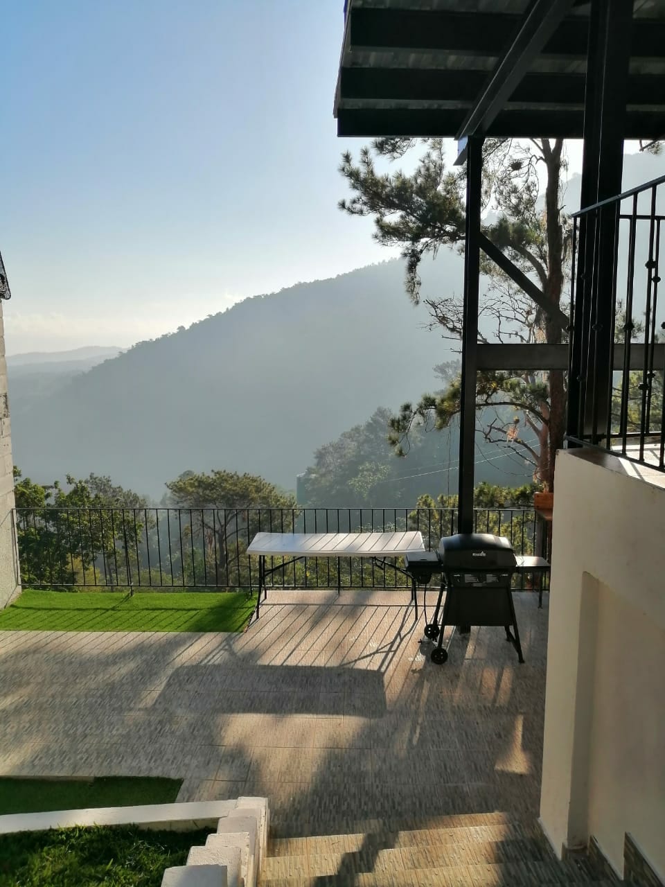 An outdoor patio area is featured, overlooking a serene mountain landscape. A charcoal grill is present next to a table, positioned on a deck with views of the greenery below. Sunlight filters through the trees, casting gentle shadows across the space.