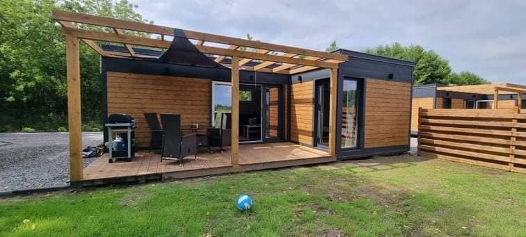 A modern cabin is shown with warm wooden cladding and sleek black accents. A spacious deck area is furnished with dark outdoor chairs and a table, while large glass doors provide access to the interior. Green grass surrounds the structure, contributing to a serene outdoor setting.