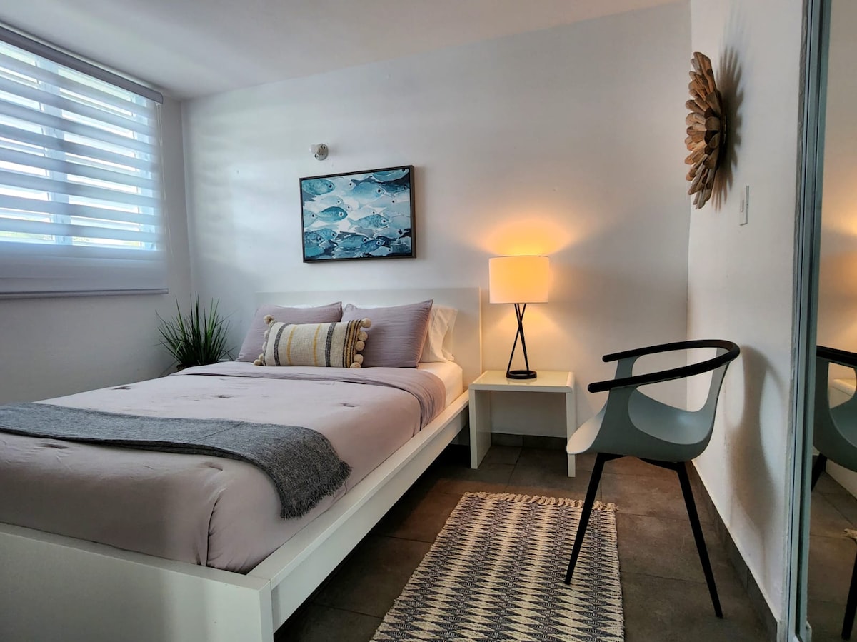 A bright bedroom features a spacious bed adorned with soft bedding. A modern lamp casts a warm light beside a minimalist nightstand. A single chair and a decorative rug complement the room's ambiance, while a piece of fish-themed wall art adds a touch of interest.