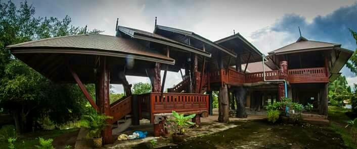 A multi-level wooden structure is showcased, featuring overhanging roofs and broad balconies. Lush greenery surrounds the building, with various plants placed throughout the base. The architecture reflects an open design, blending seamlessly with the natural environment.