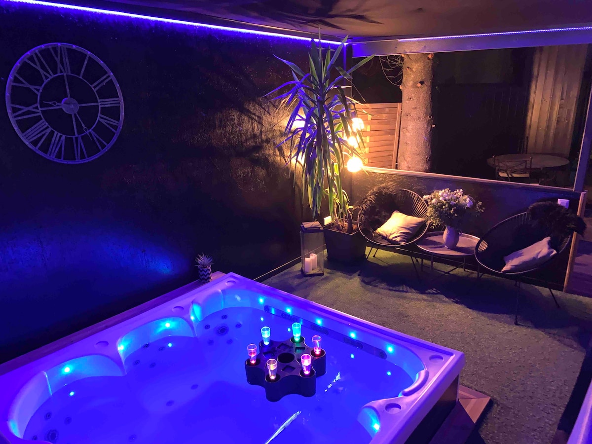 An outdoor jacuzzi is highlighted by soft blue lighting, surrounded by lush greenery. Comfy seating is arranged nearby with decorative cushions, and candles sit atop the jacuzzi, enhancing the tranquil ambiance. A clock adorns the wall, ensuring a sense of time in this relaxing space.