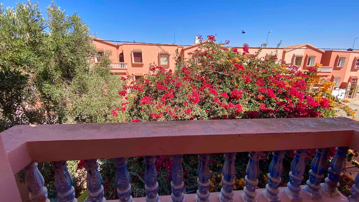 A view from a balcony is presented, overlooking vibrant pink bougainvillea that envelops the garden. The picturesque surrounding buildings feature warm-colored facades under a clear blue sky, enhancing the outdoor ambiance.