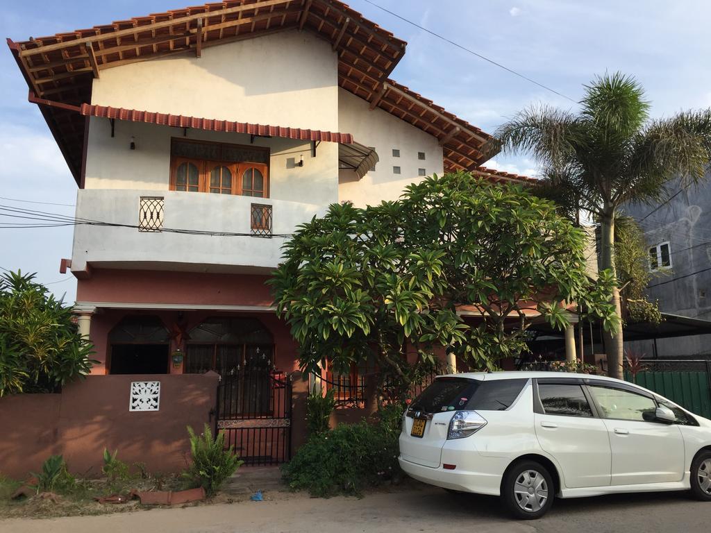 A two-story villa is shown, featuring a combination of white and red exterior walls. A roof with wooden beams extends over the upper balconies. Lush green plants frame the entrance, while a parked white vehicle is visible in the foreground, indicating available parking.