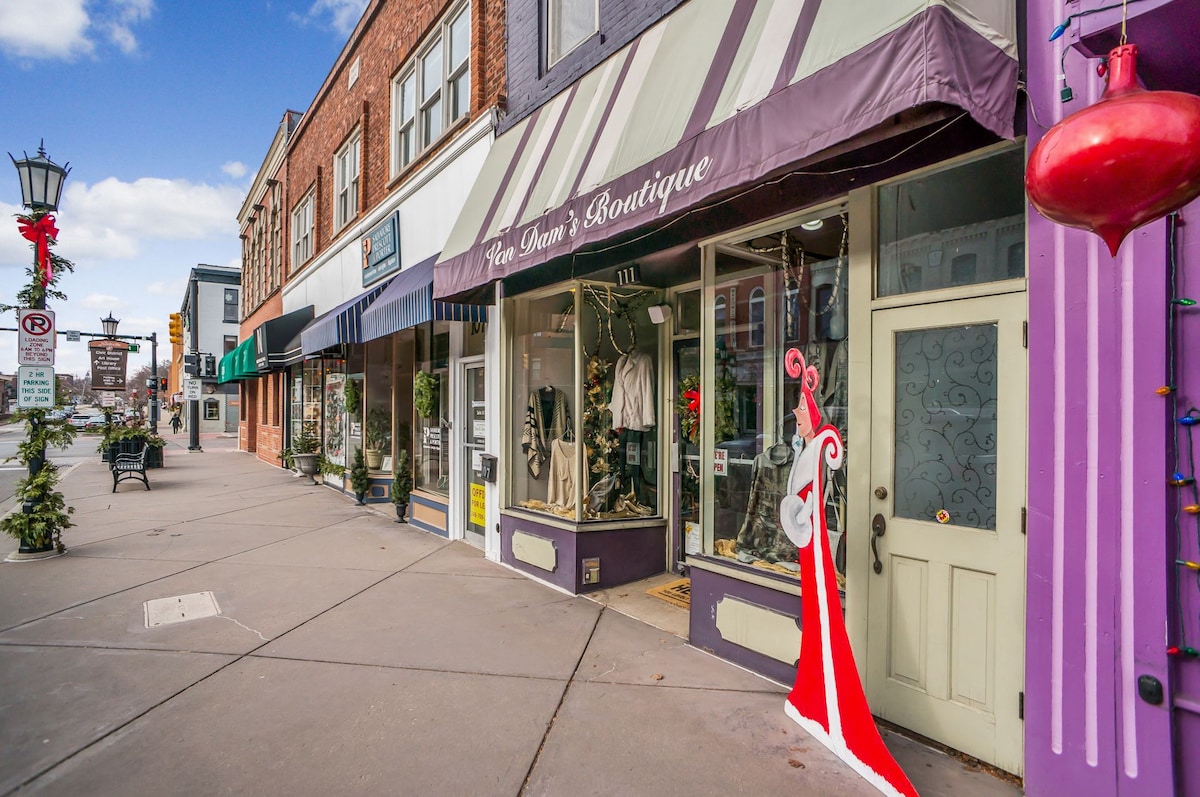 A charming street view of historic downtown Northville showcases a collection of boutique shops with colorful awnings. Festive decorations add a seasonal touch, while a wide sidewalk invites leisurely strolls. The scene captures the vibrant atmosphere of the area, emphasizing the accessibility to shops and dining options.