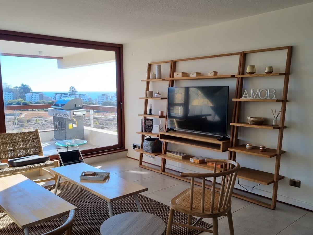 A living area is showcased with a wooden bookshelf displaying decorative items and a television. Two chairs accompany a low coffee table, and a textured rug is laid on the tile floor. Large windows offer a glimpse of the sea view and outdoor barbecue area.
