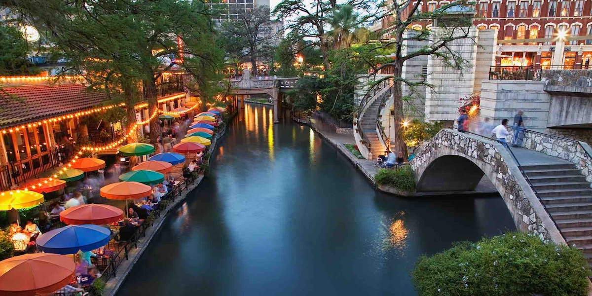 The image captures a serene section of the River Walk, featuring colorful umbrellas shading outdoor seating along the water. Soft lights illuminate the venue, and lush trees line the banks. A graceful stone bridge connects the two sides, enhancing the scenic view.