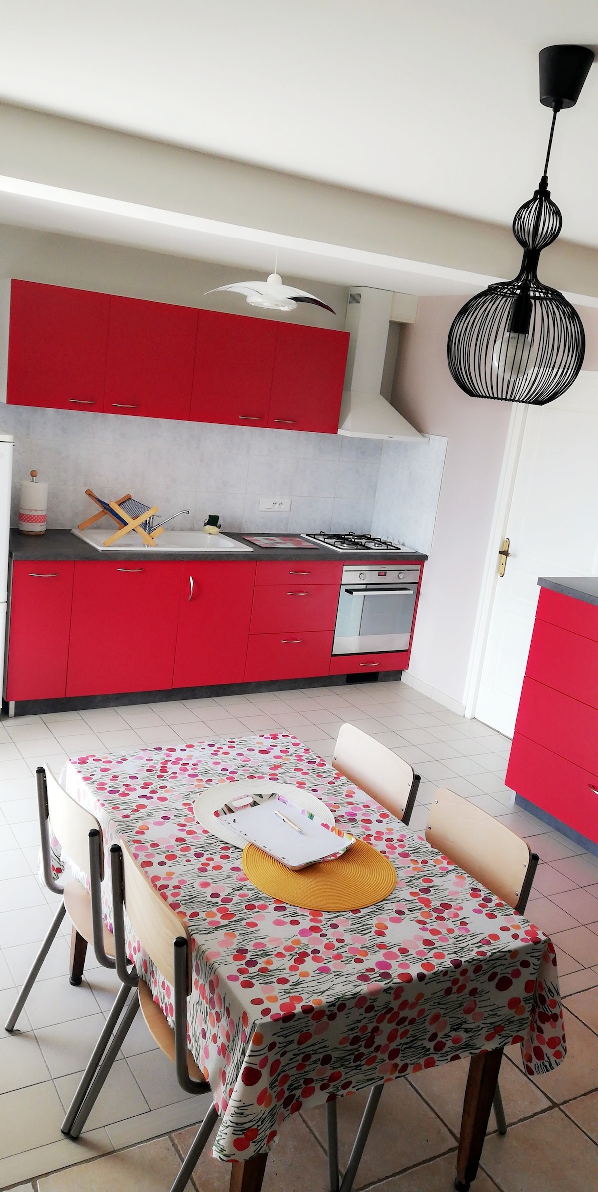 A modern kitchen features red cabinetry and a light-colored countertop. A dining table with a floral tablecloth accommodates four chairs. Above, a unique pendant light adds character to the space. Appliances include an oven, stovetop, and an extractor hood, enhancing functionality.