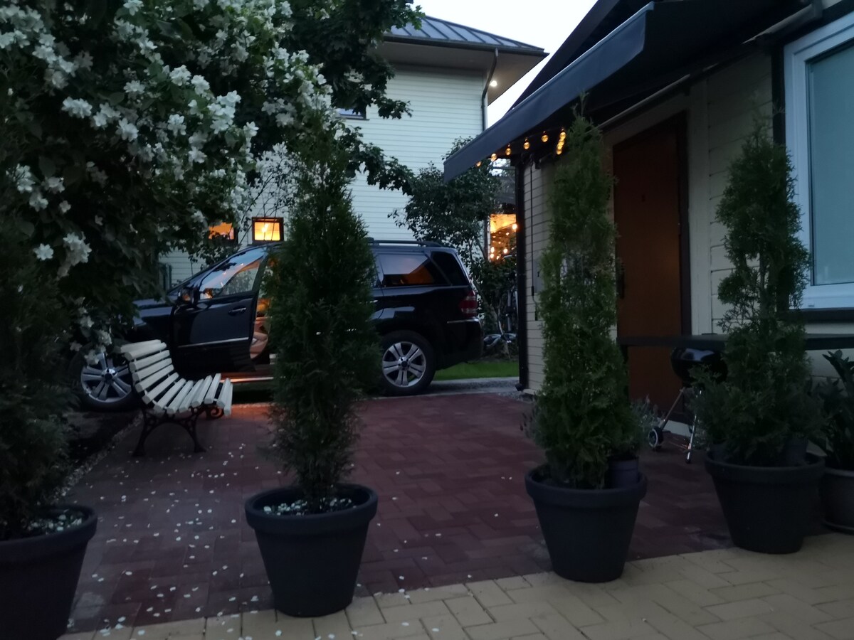 An outdoor area is depicted, featuring potted shrubs and a vehicle parked nearby. The warm glow of interior lights is visible through windows, suggesting a welcoming atmosphere. A chair and a bicycle are also present within the space, surrounded by blooming flowers.