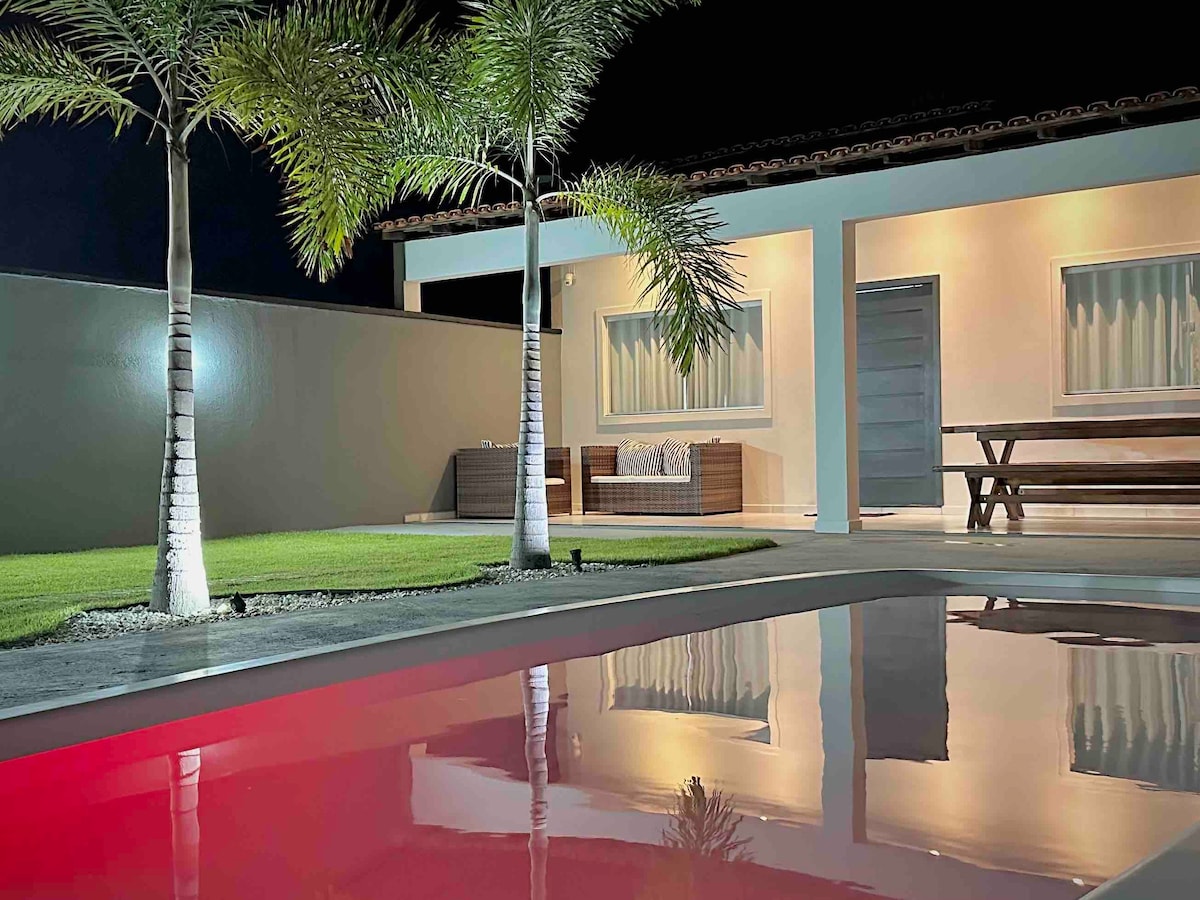 A tranquil outdoor area features a private pool reflecting soft red hues under night lighting. Palm trees are situated beside the pool, enhancing the serene ambiance. A shaded seating arrangement is visible in the background, complemented by a well-manicured grassy area.