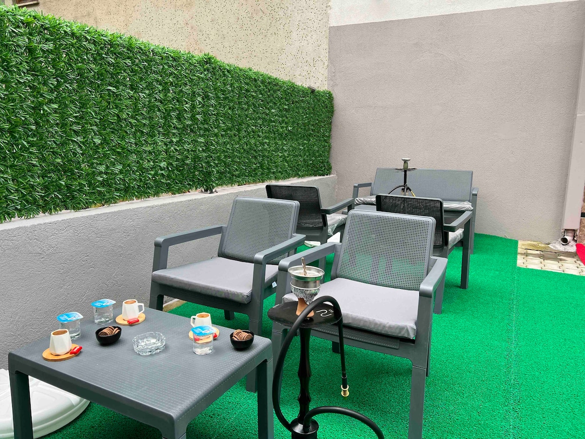 An outdoor seating area features several grey chairs with soft cushions arranged around low tables. Green artificial grass covers the ground, and decorative greenery lines the wall. Several small cups and an ashtray are visible on the tables, along with a hookah.