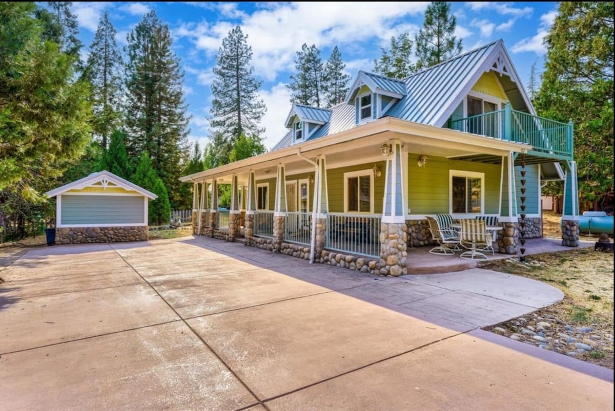 A two-story A-frame house is depicted with a light yellow exterior and green balcony. The front porch is furnished with multiple seating options. A spacious driveway leads to a nearby outbuilding. Surrounding pine trees provide natural shade, enhancing the serene ambiance.