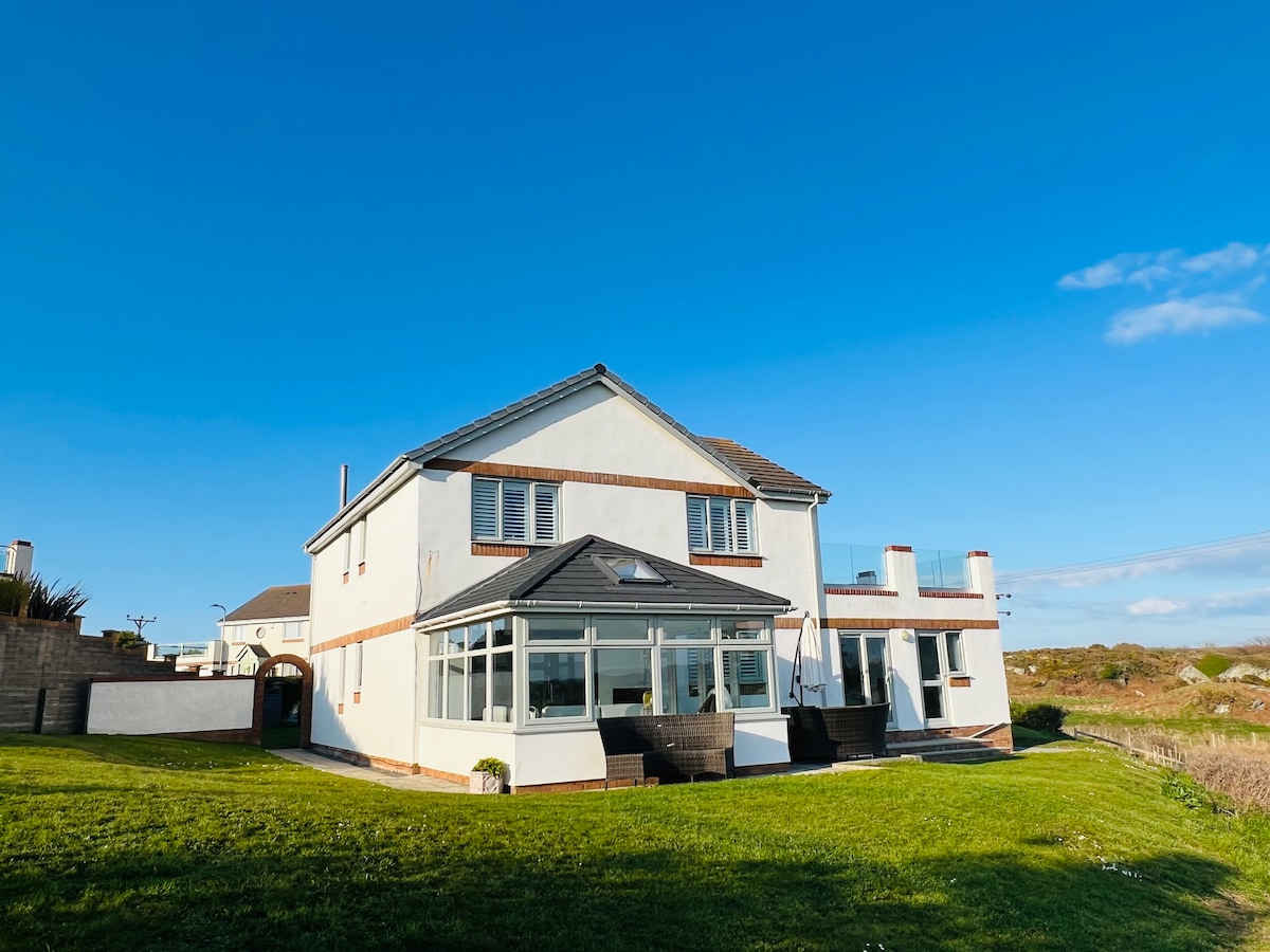 A detached house is presented against a clear blue sky, showcasing a well-maintained lawn and expansive garden. Large windows reflect natural light, and a sunroom enhances the open feeling. The structure features both a welcoming facade and a spacious outdoor area for relaxation.