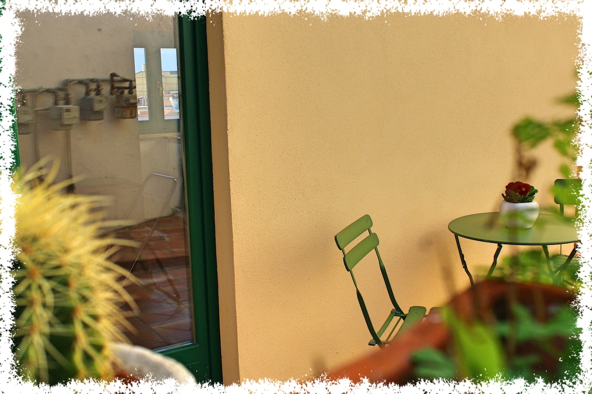 A small outdoor seating area is visible, featuring a round table and two green chairs. The wall behind is painted a neutral tone, while a potted plant adds a touch of greenery. A glimpse of the interior space can be seen through a glass door.