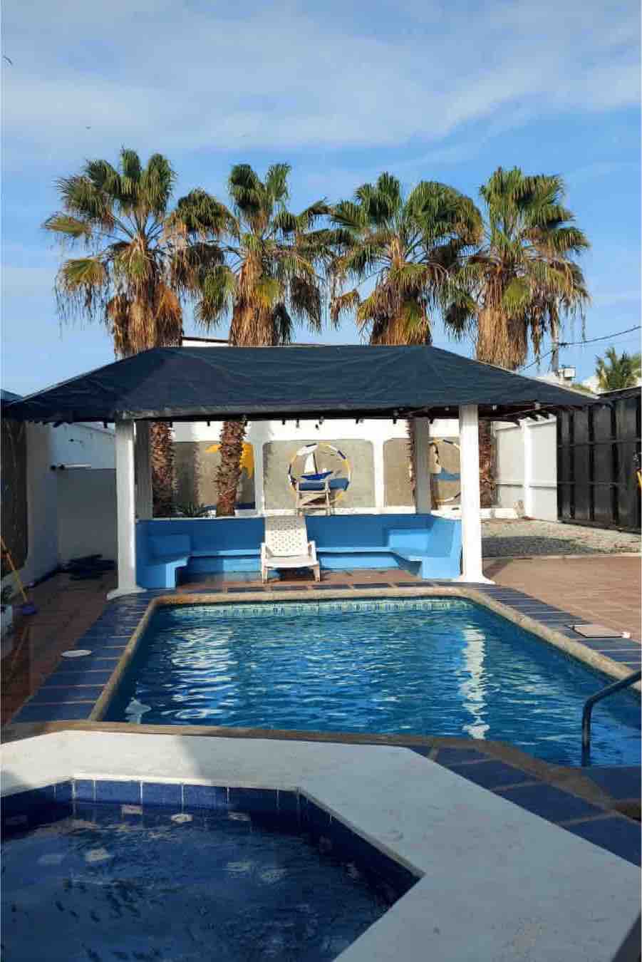 A private pool area features a rectangular pool with a surrounding paved deck, a shaded gazebo, and multiple palm trees. A single white lounge chair rests under the gazebo, providing a relaxing spot. The backdrop includes a wall and a sunny sky.