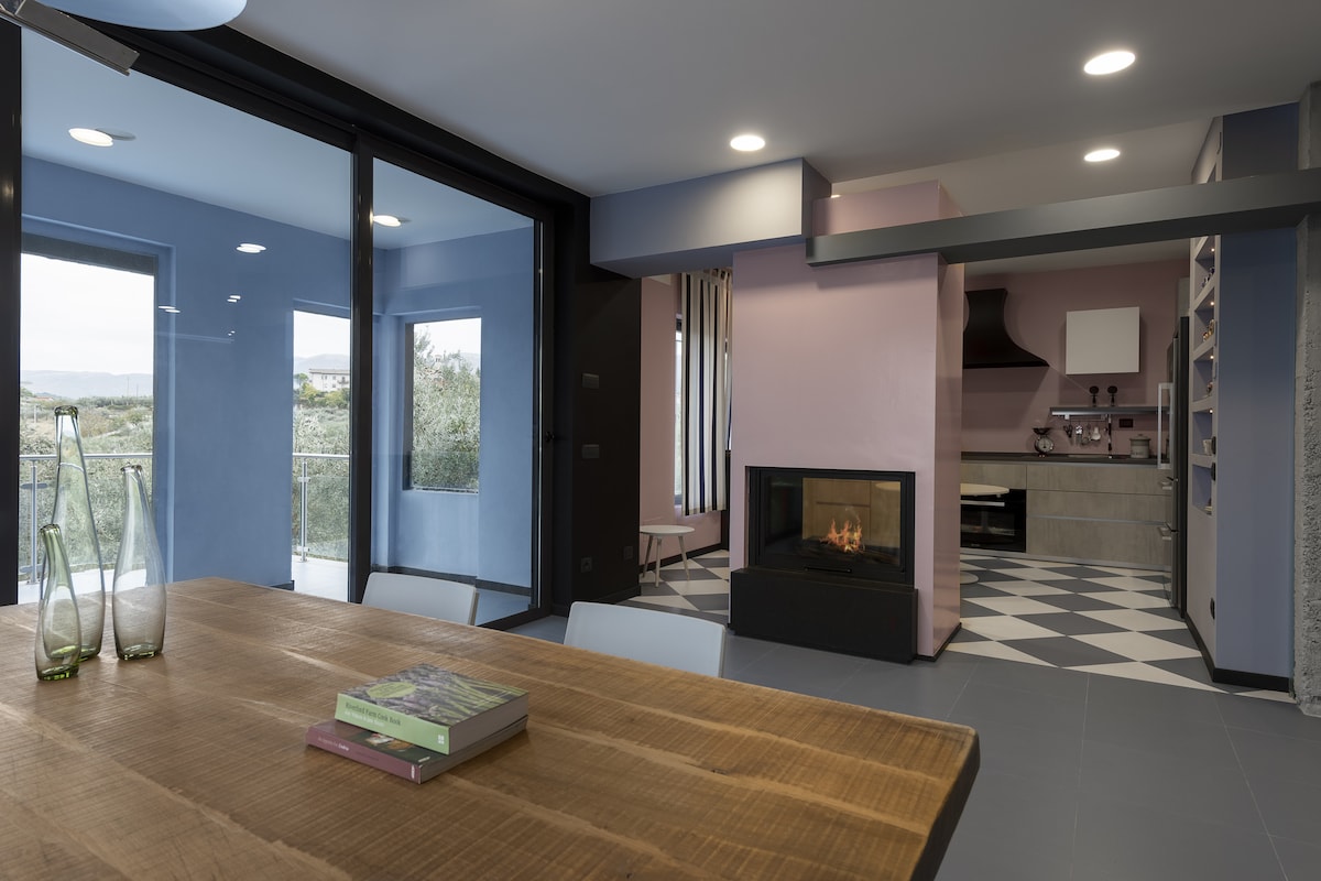 A spacious dining area features a large wooden table, complemented by minimalist decor. A modern fireplace is set against a wall in soft pink, while large glass doors open to a scenic outdoor view, enhancing the light within the room.