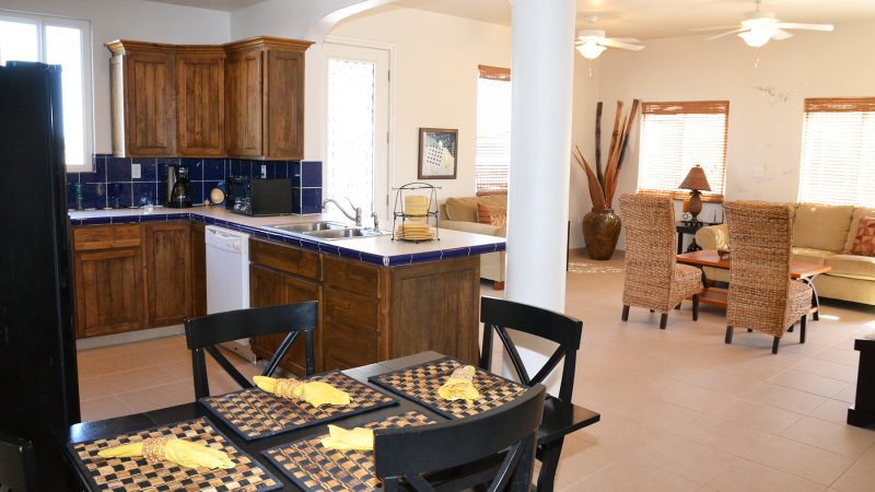 An open-concept kitchen and living area are visible, featuring wood cabinets, a blue-tiled backsplash, and a dining table set for two. Comfortable seating is arranged in the living area, accompanied by decor elements like potted plants and woven textures.