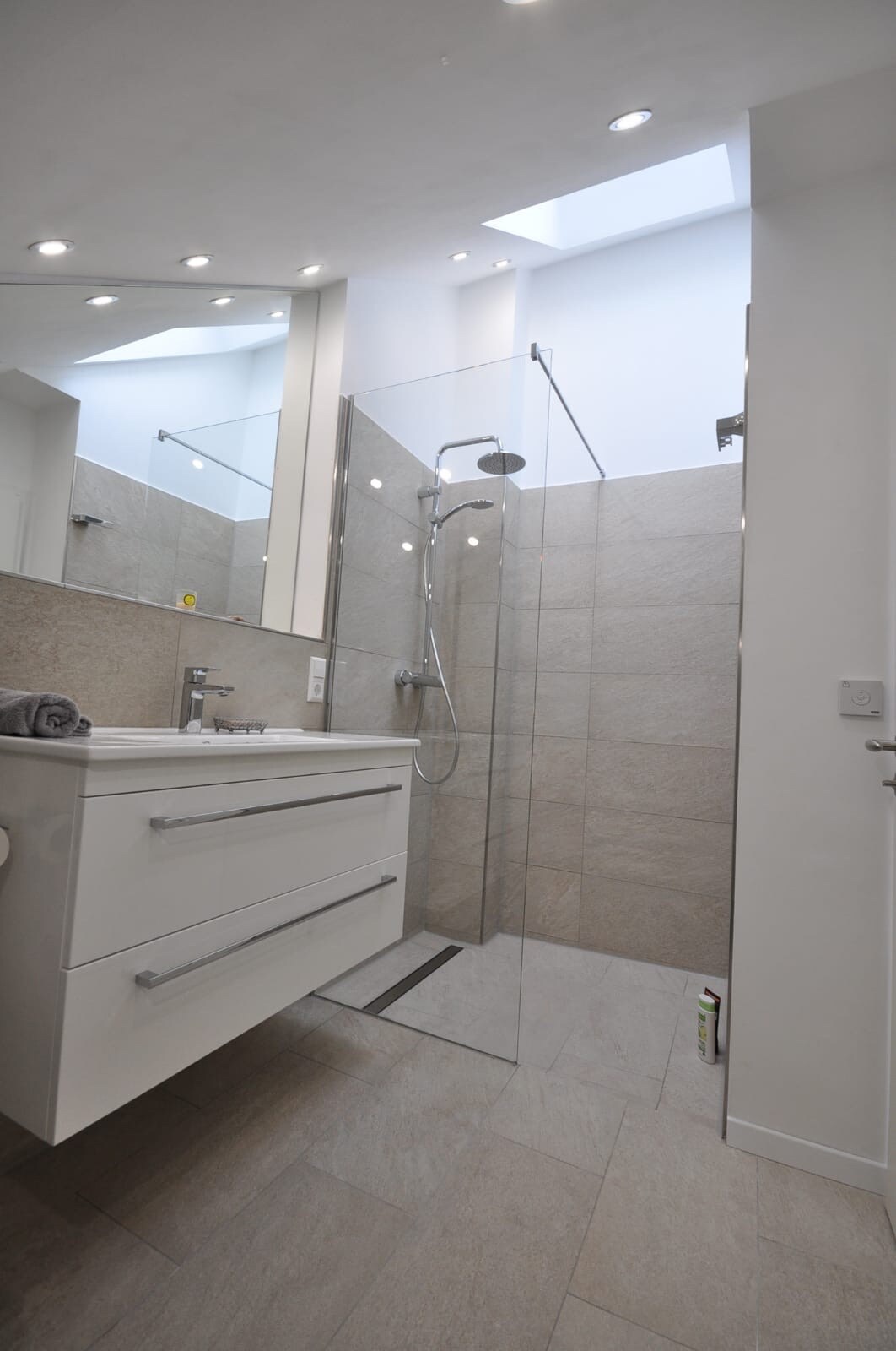 A modern bathroom features a walk-in shower with a glass enclosure and overhead rain showerhead. Sleek white cabinetry complements the light-toned tile flooring, while natural light floods in from a skylight above. A minimalist design is emphasized by clean lines and a spacious feel.