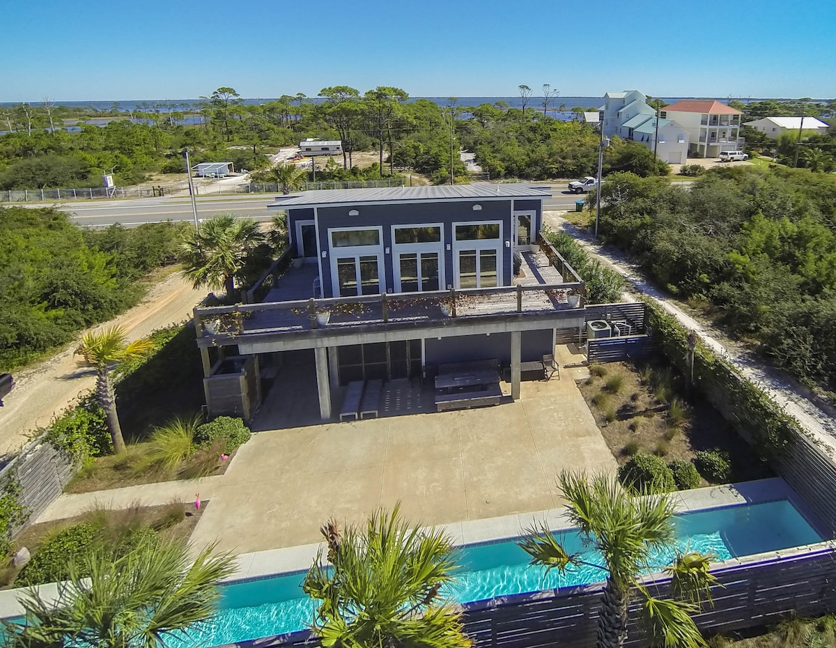 A modern coastal retreat is shown from an aerial perspective, featuring a fenced lap pool bordered by palm trees. The outdoor space includes a patio with seating options. In the background, the Gulf of Mexico and surrounding greenery are visible, framed by a clear blue sky.