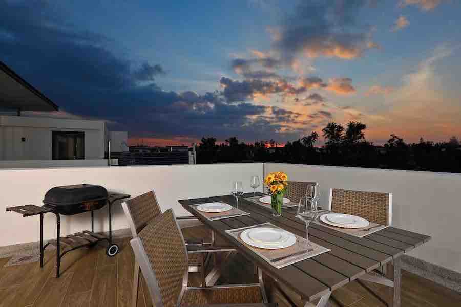 An outdoor dining area features a rectangular table surrounded by six wicker chairs. A grill is positioned nearby. A vase with a bouquet of flowers is centered on the table alongside table settings. The sunset sky casts warm colors across the scene.