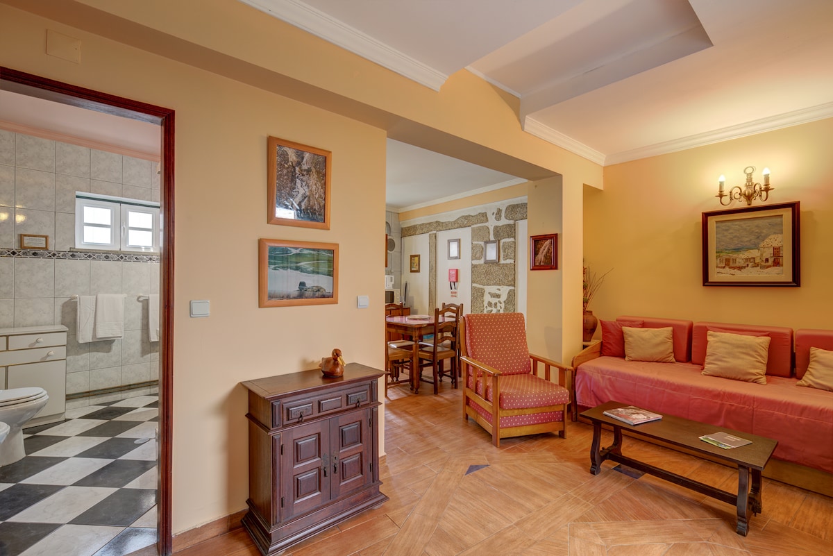 A welcoming living area features a red sofa and armchair, complemented by neutral walls. Decorative artwork enhances the space, while a wooden table adds functionality. The adjoining room showcases a bathroom with tiled walls, and natural light enters through multiple windows, creating an open and airy ambiance.