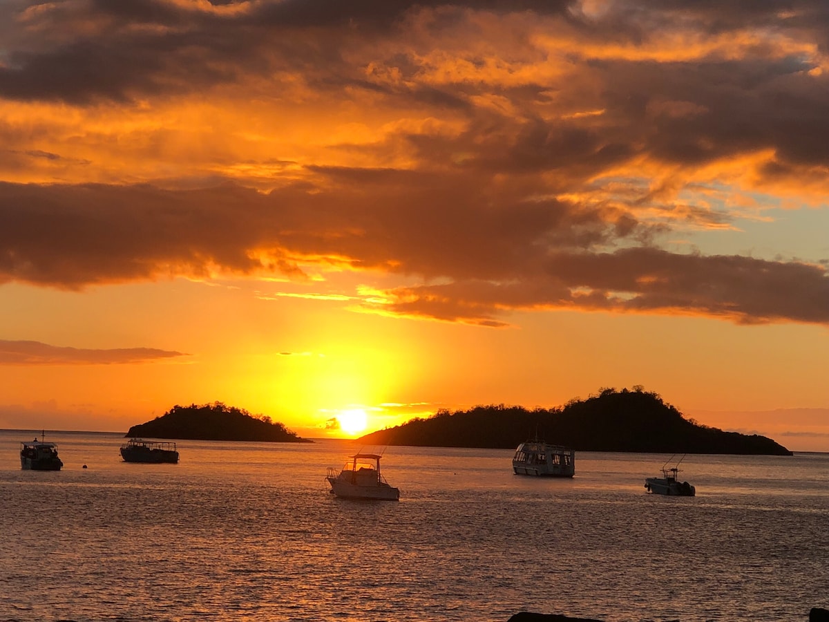 A vibrant sunset casts warm orange and yellow hues across the water, illuminating several boats anchored in the foreground. Silhouettes of distant islands are visible against the colorful sky, creating a serene coastal scene.
