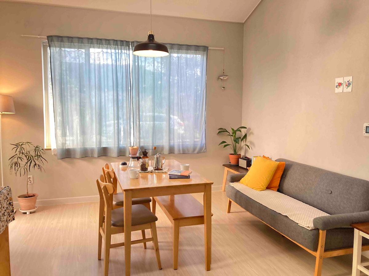 A dining area is presented with a wooden table and four chairs, positioned under a pendant light. A gray sofa with an accent pillow is situated nearby. Large windows are dressed with light curtains, allowing natural light to fill the space. A small indoor plant adds a touch of greenery.
