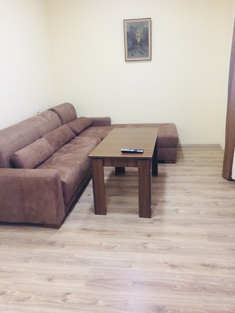 A comfortable living area is furnished with a brown fabric sofa and a wooden coffee table. A remote control rests on the table, and light wooden flooring complements the pale yellow walls. A framed artwork adds a subtle touch to the room.