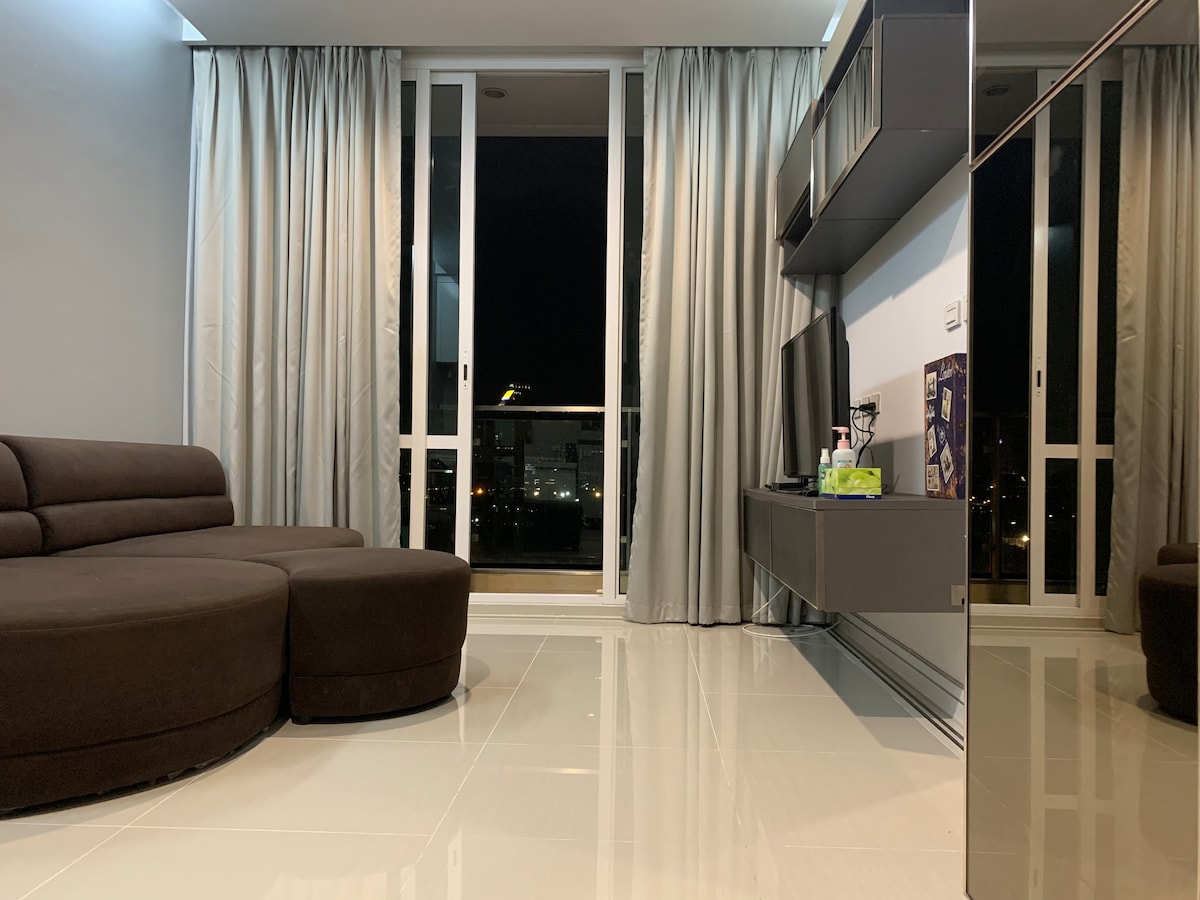 A modern living area is displayed, featuring a circular brown sectional sofa positioned against a wall. Large glass doors offer a view to the outside, framed by light gray curtains. A wall-mounted TV and sleek cabinetry complement the space, reflecting a polished tile floor.
