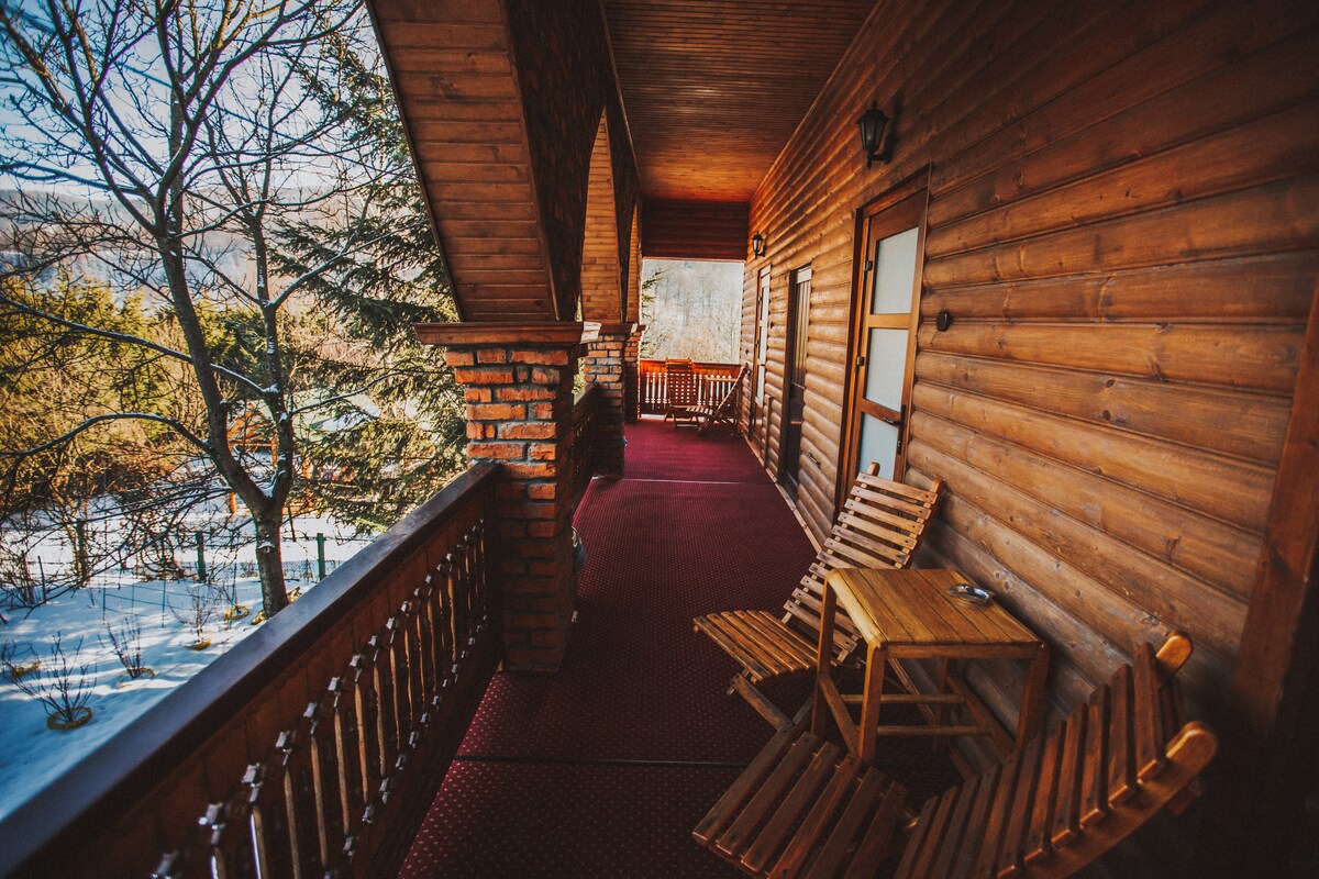 An outdoor balcony is seen, featuring wooden flooring and a covered area. Two wooden lounge chairs are positioned beside a small table, inviting relaxation. The wooden railing and columns enhance the natural ambiance, while trees and a snowy landscape are visible in the background.