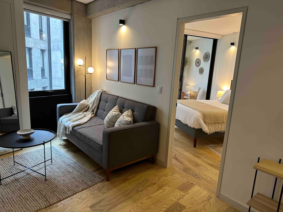 A modern living room features a gray sofa adorned with light-colored cushions, accompanied by a round black coffee table. Soft lighting from wall sconces and natural light enters through large windows. A doorway leads to a bedroom area, visible in the background.