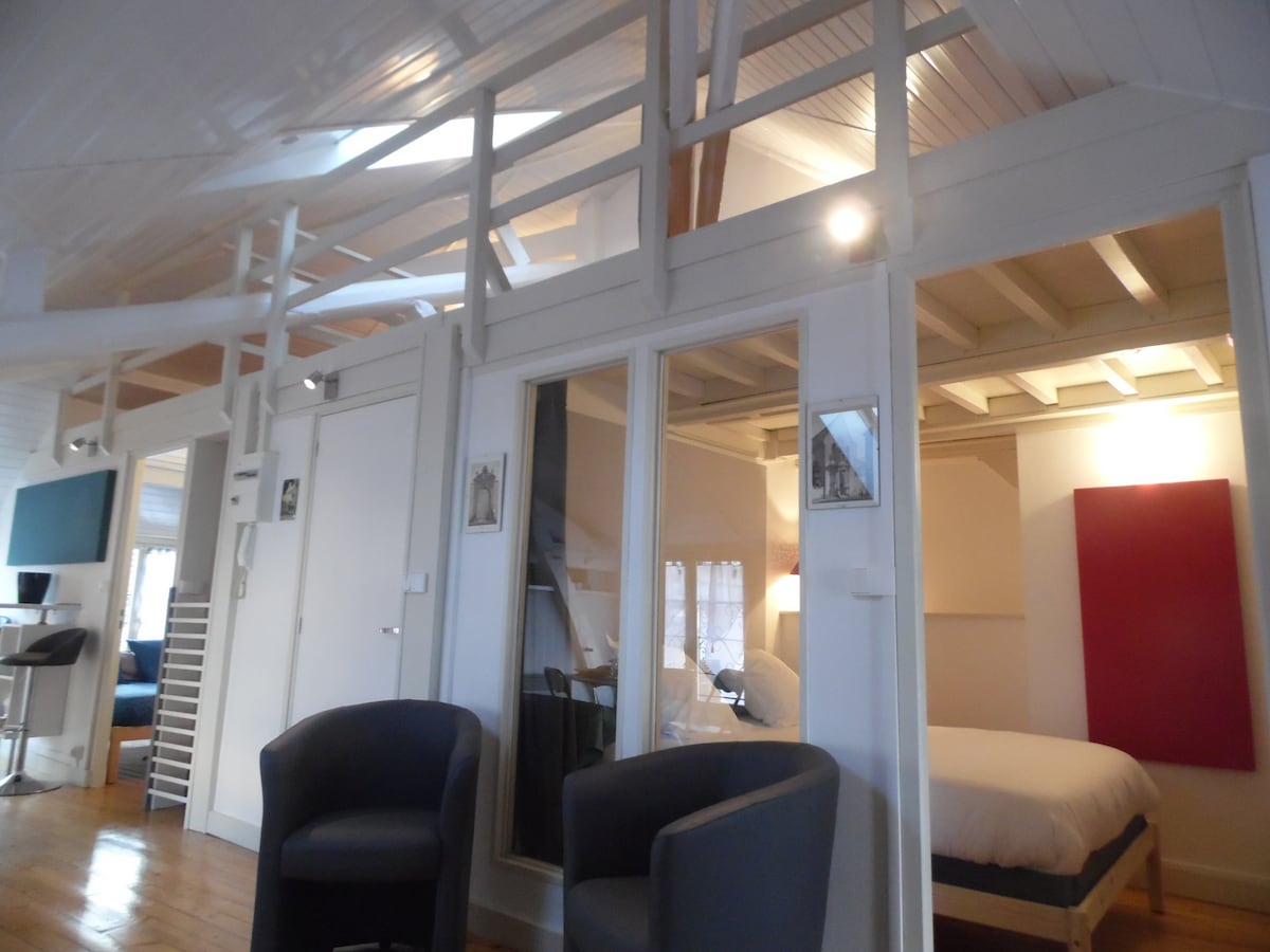 The image showcases a bright, open space with wooden beams and a cozy atmosphere. Two comfortable armchairs are placed in the foreground. In the background, a double bed is visible, along with a workspace featuring a small desk. Natural light filters through the windows.