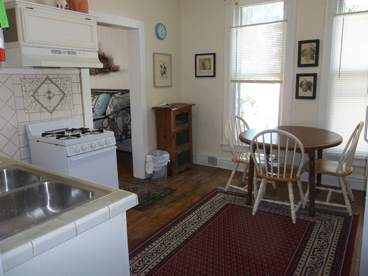 A bright kitchen area features a gas stove and a double sink, with light streaming through a nearby window. A wooden dining table with three chairs is positioned beside a rustic cabinet. Decorative wall art and a rug add warmth to the space.