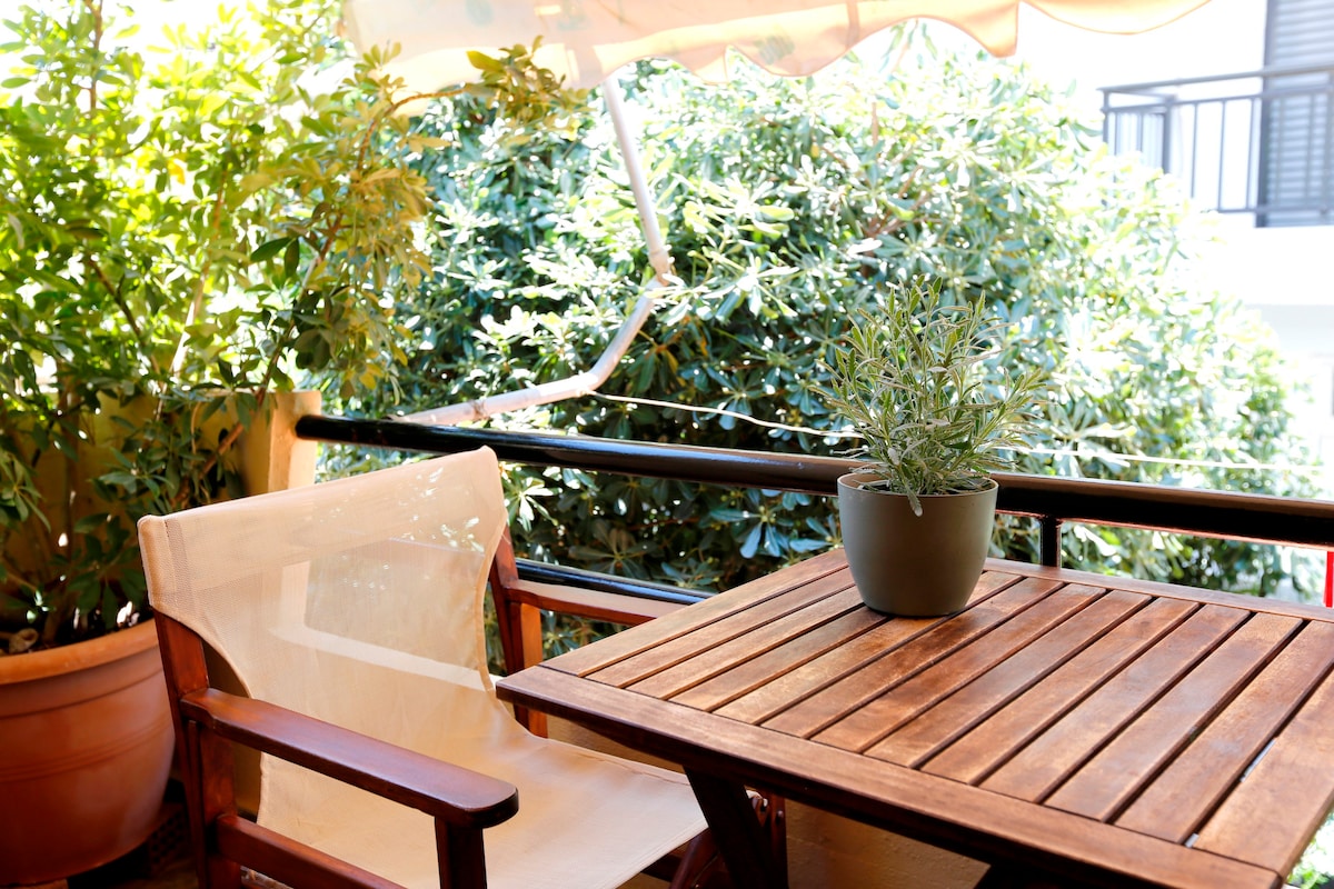 A private balcony features a wooden table accompanied by a comfortable chair. A potted plant adds a touch of greenery, while leafy bushes provide natural privacy. The awning offers shade, contributing to a peaceful outdoor setting.