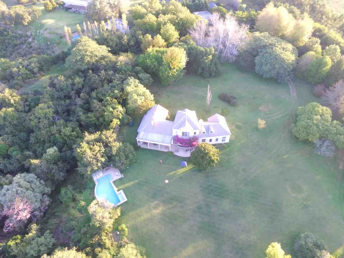 An aerial view captures the expansive property with lush greenery surrounding a large house. A glistening pool is positioned adjacent to the residence amidst beautifully landscaped gardens, providing a serene ambiance. The vast open space invites exploration, highlighted by the greenery of the indigenous forest.