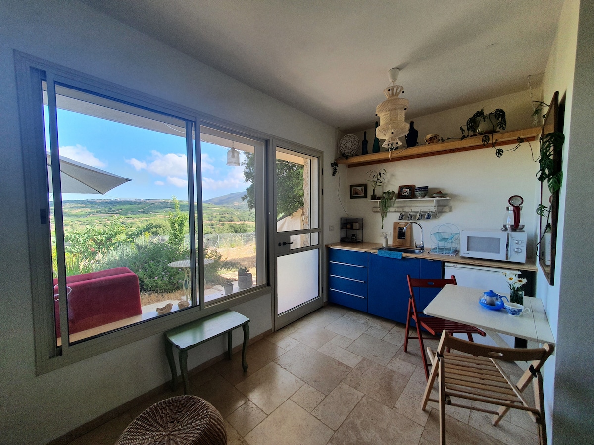 A light-filled kitchenette features a blue cabinet and a microwave. A small dining table with two chairs is positioned next to the large window, providing views of the green landscape outside. Natural light enhances the stone floor and warm textures within the space.