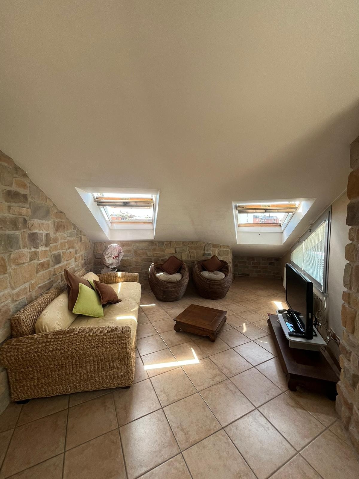 A cozy sitting area features a woven sofa adorned with green and brown cushions. Two comfortable bean bag chairs are positioned nearby. A wooden coffee table sits on tiled floors, with natural light streaming through two skylights that brighten the stone walls.